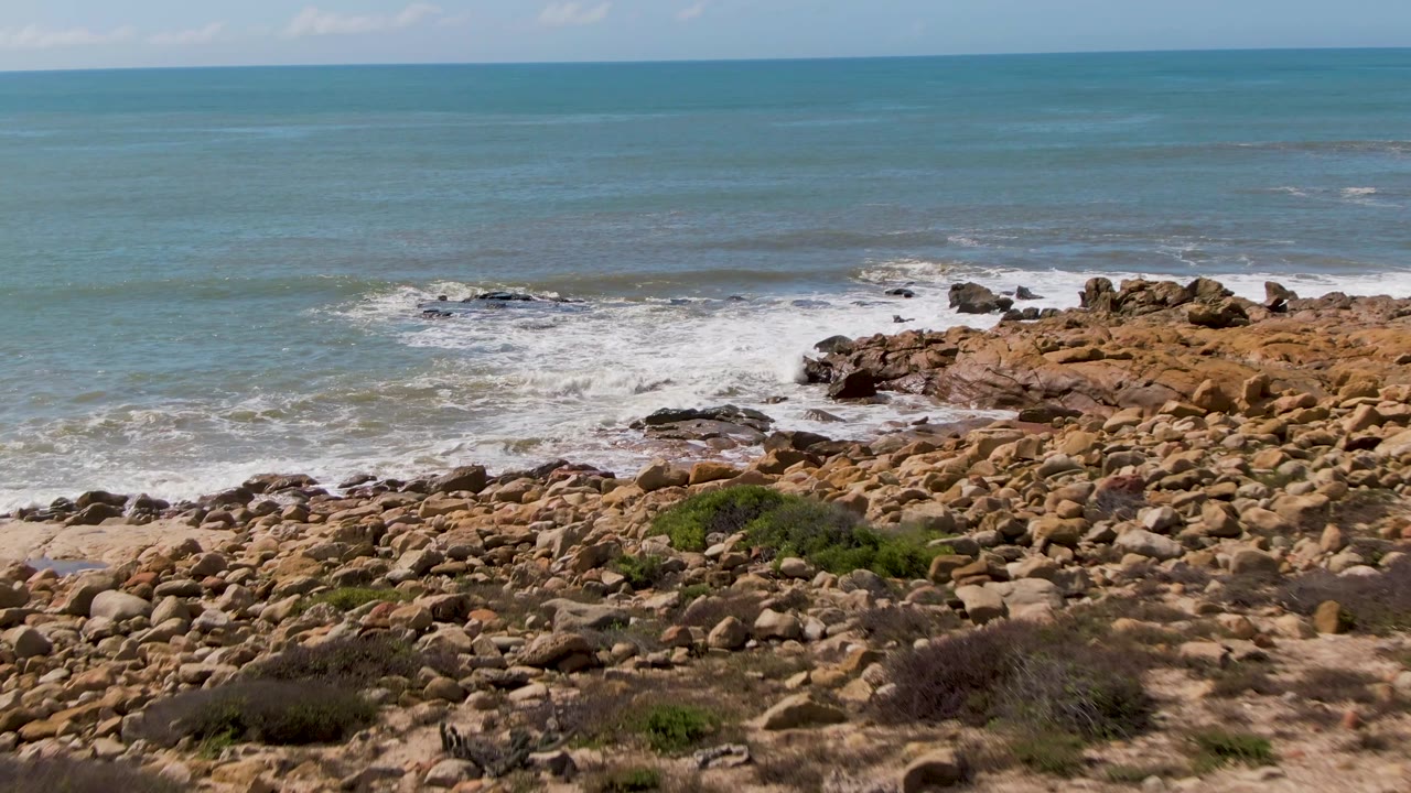 At 0:02, the rocky point at Buzzards Beach takes more of the right side of frame as white foam collects along the shore, with open blue-green water behind it and no visible people.