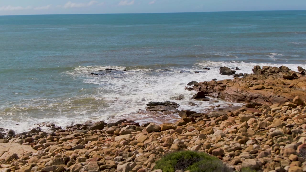 At 0:03, the frame is tighter on the surf zone at Buzzards Beach, with rounded tan rocks across the lower half, white water on the left, ocean beyond, and no visible people.