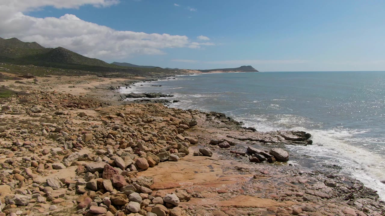 Wide elevated view of Buzzards Beach East Cape in Mexico with tan boulders in the foreground, waves breaking on dark rock shelves, distant mountains, and no visible people.