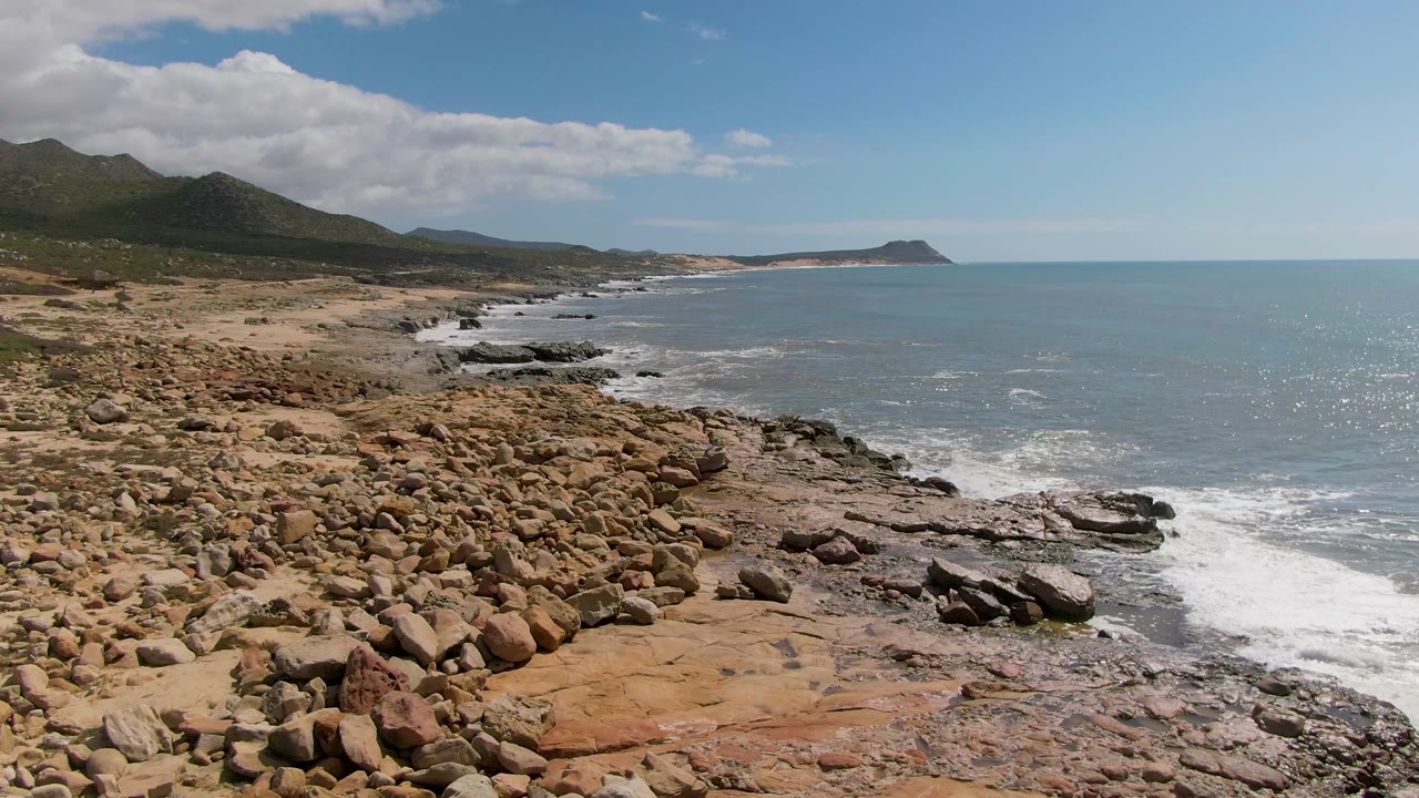 Buzzards Beach East Cape coastline seen from above with rust-colored rocks in front, whitewater spreading over the shore rocks, a curved bay, distant headland, and no visible people.