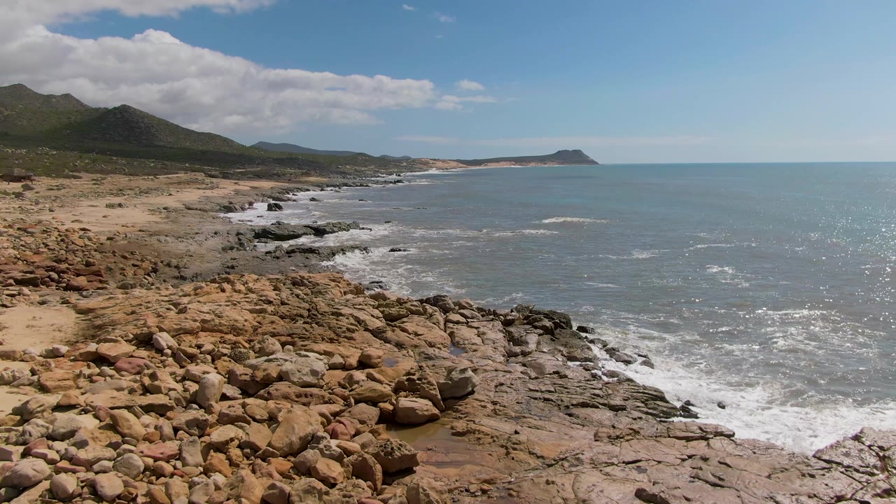 Opening view of Buzzards Beach, East Cape, BCS Mexico, with brown rocks in the foreground, surf breaking along the shore, distant hills at left, and no visible people.