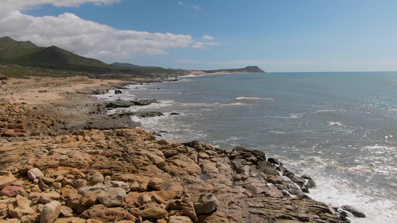 Buzzards Beach coastline seen from above with jagged foreground rocks, white foam wrapping the shoreline, a sandy strip beyond, and no visible people.
