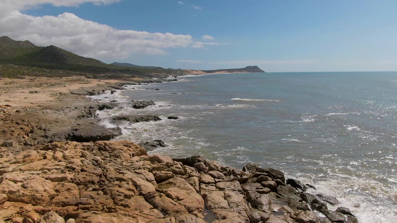 A slightly later view of the Buzzards Beach shore shows more foreground rock texture, smaller foam patches along the coast, the distant headland, and no visible people.