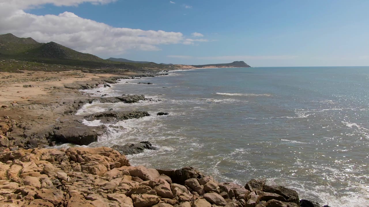 Waves strike a rocky cove below dry hills and a partly cloudy sky.