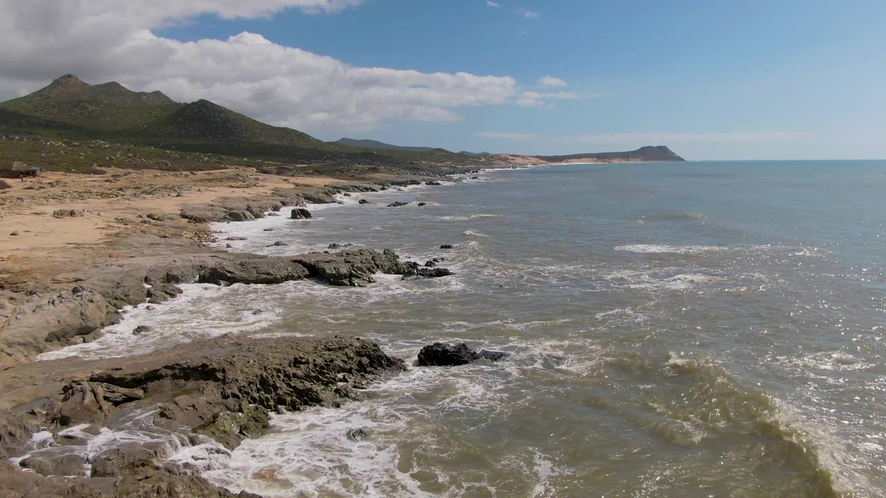 Buzzards Beach East Cape BCS Mexico shoreline with darker rocks closer to camera, active whitewater along the beach, open blue-green sea to the right, and no visible people.