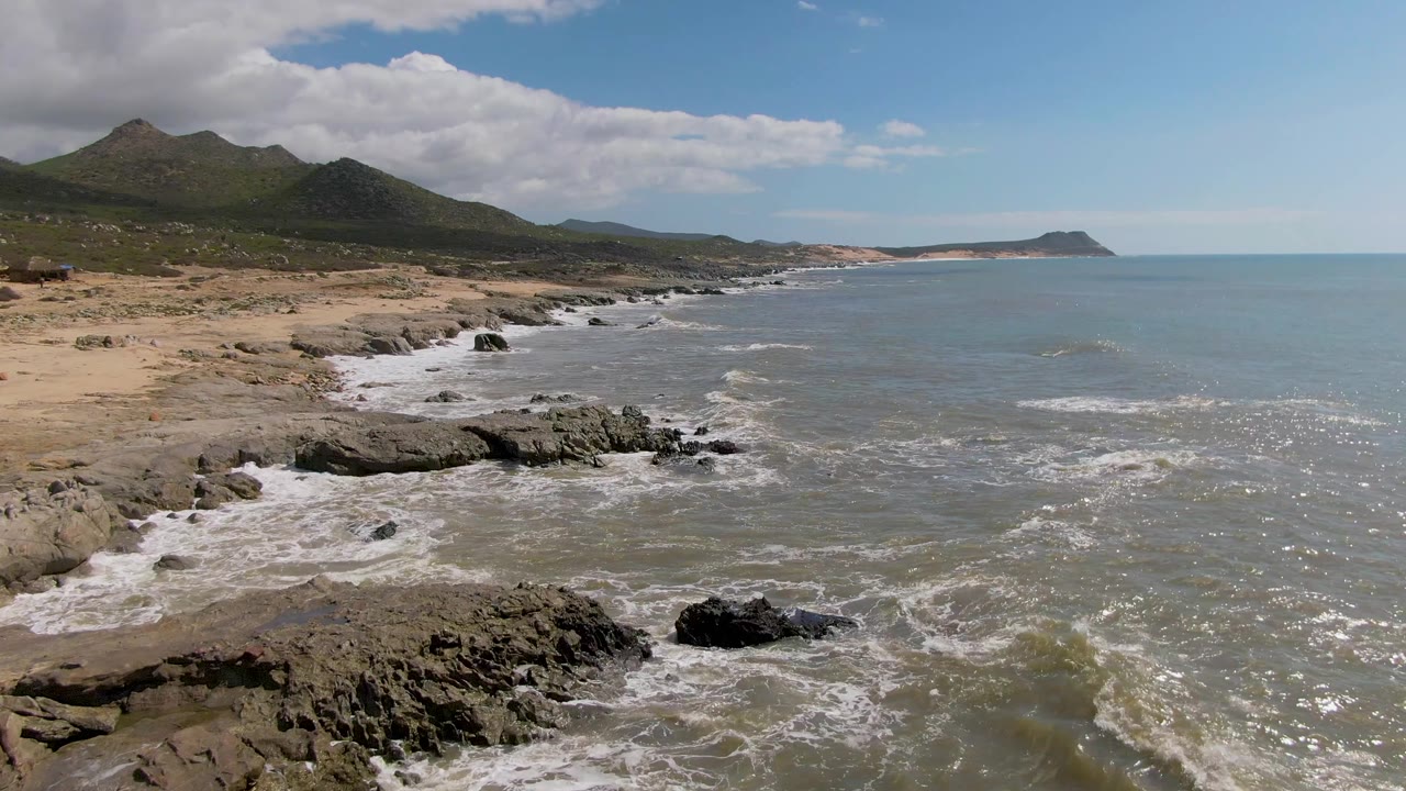 Elevated view over Buzzards Beach East Cape BCS Mexico with a rocky ledge in the lower frame, foam rolling along the shore, broad ocean on the right, mountains inland, and no visible people.