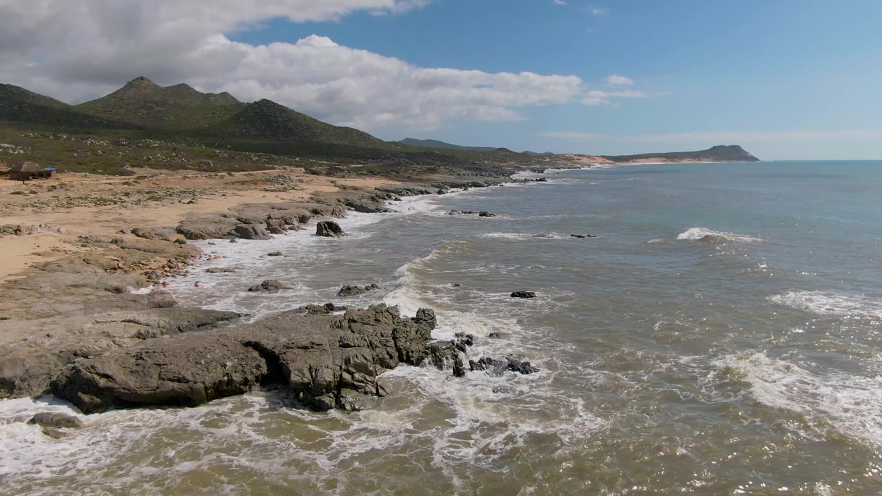 Opening frame of Buzzards Beach East Cape BCS Mexico showing white surf around foreground rocks, a sandy shoreline to the left, green hills behind it, and no visible people.