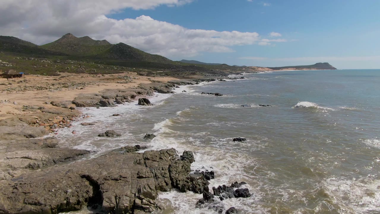Buzzards Beach East Cape BCS Mexico from a high coastal view, with foamy waves spreading over rock shelves, pale sand along the left edge, and no visible people.