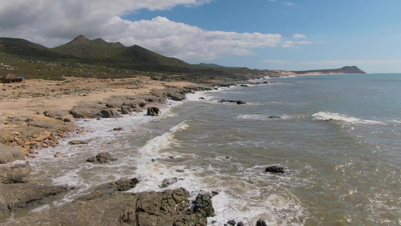 Mid-clip view of Buzzards Beach East Cape BCS Mexico showing surf pulling around scattered rocks, a broad ocean surface to the right, green hills to the left, and no visible people.