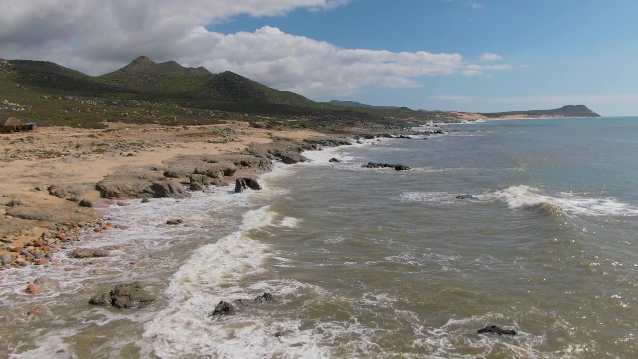 Later frame at Buzzards Beach East Cape BCS Mexico with the sandy beach more prominent, surf bands pushing into the lower left, mountains under a cloud shadow, and no visible people.