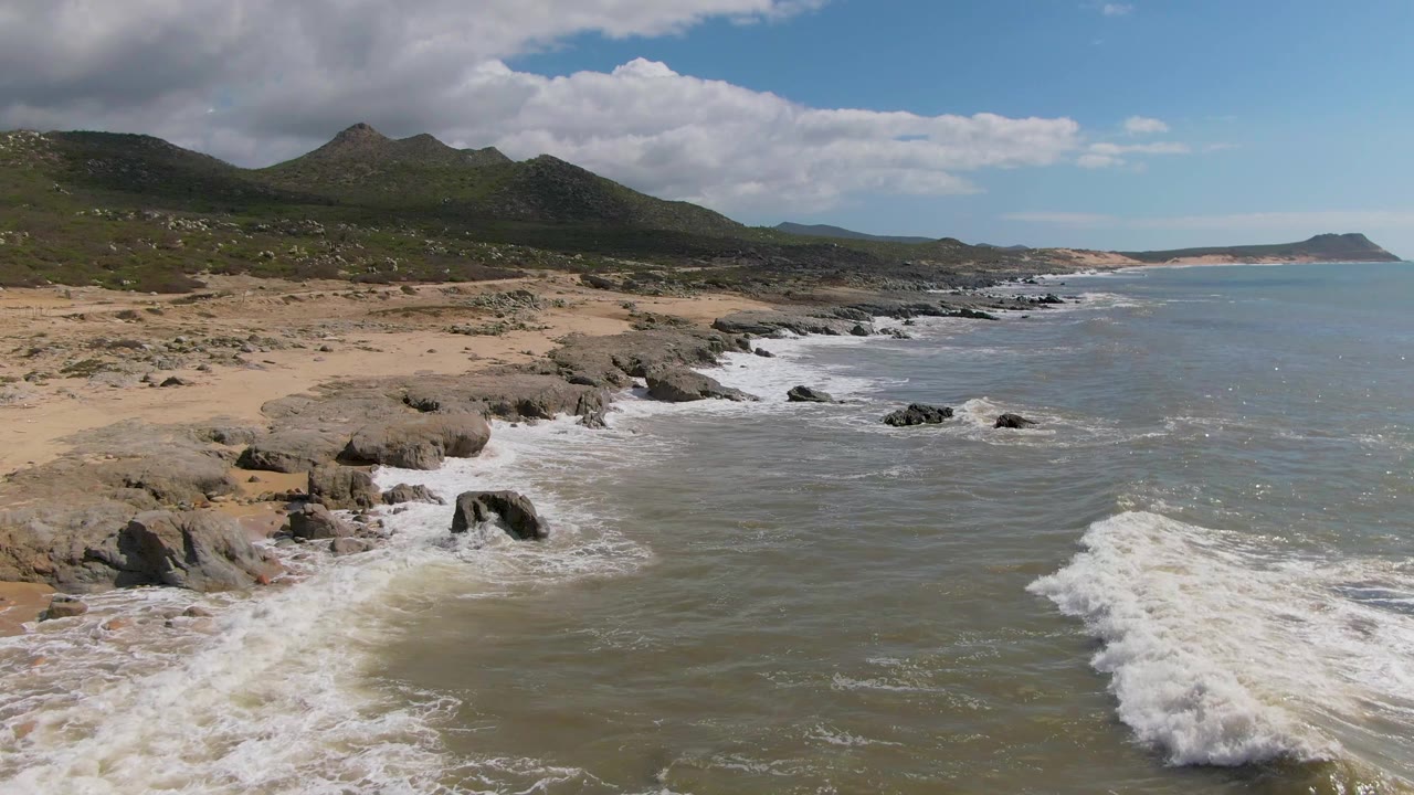 Opening frame at Buzzards Beach East Cape BCS Mexico, showing foamy surf rolling onto dark shoreline rocks, tan sand, green mountains, and no visible people.