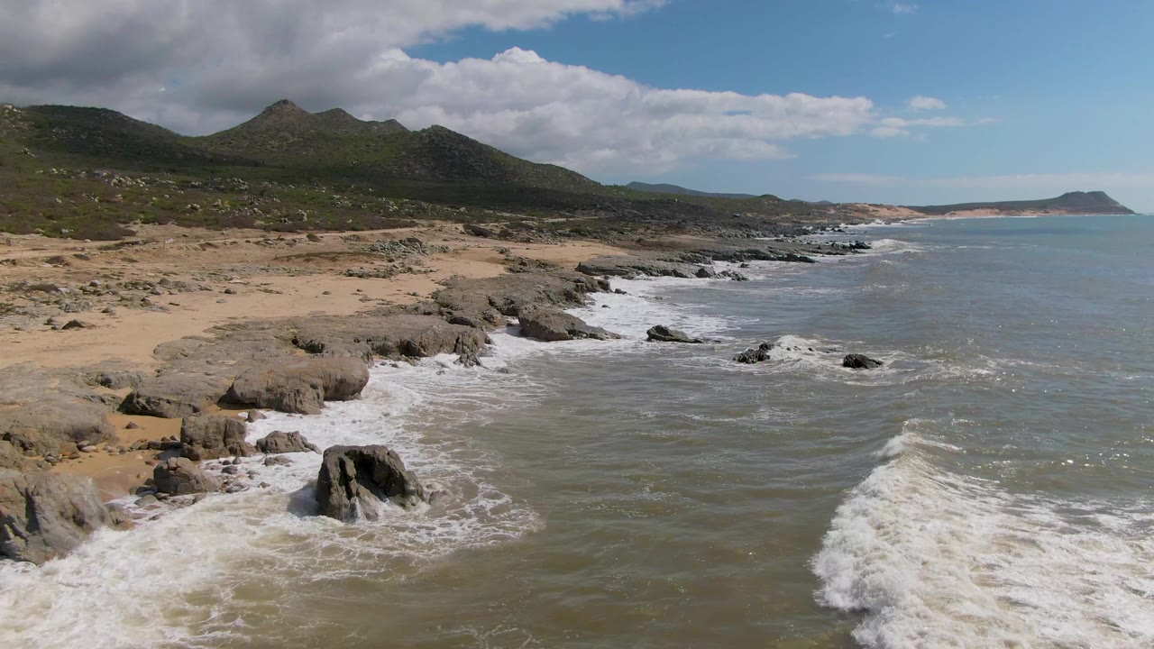 One second into the clip, a white wave line spreads around the near rocks while the sandy Buzzards Beach shoreline and mountain ridge remain clear, with no visible people.