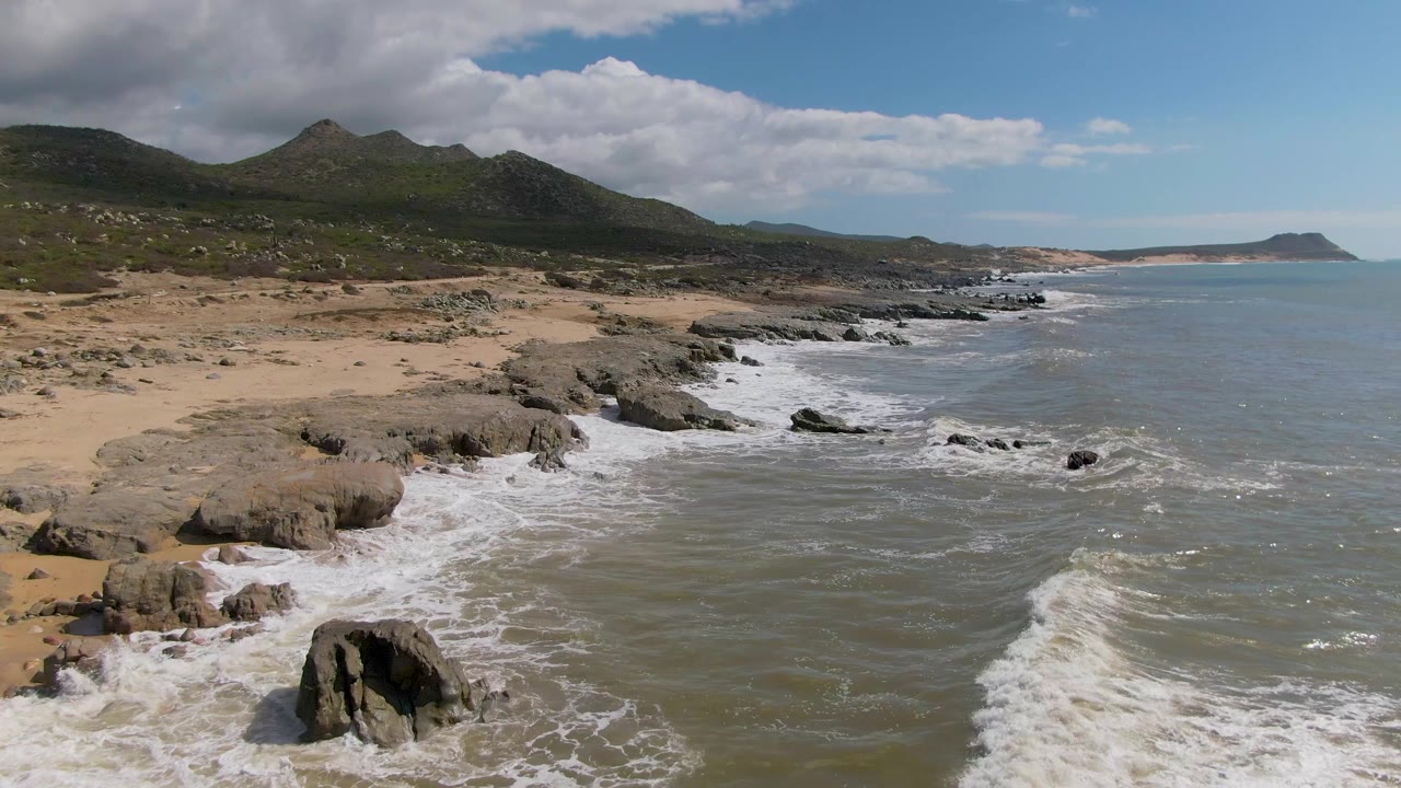 At 0:02, a prominent dark rock sits near the lower center as surf curls around it, with Buzzards Beach sand, coastal rocks, mountains, and no visible people in view.