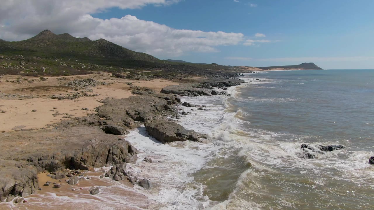 First frame of Buzzards Beach East Cape BCS Mexico showing white surf washing around dark rocks, tan sand, green hills, blue ocean, partly cloudy sky, and no visible people.