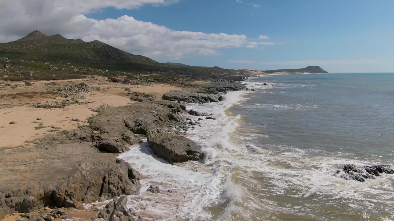 Buzzards Beach East Cape BCS Mexico frame with foamy water pushing farther into the rocky foreground, tan beach to the left, distant hills, ocean horizon, and no visible people.