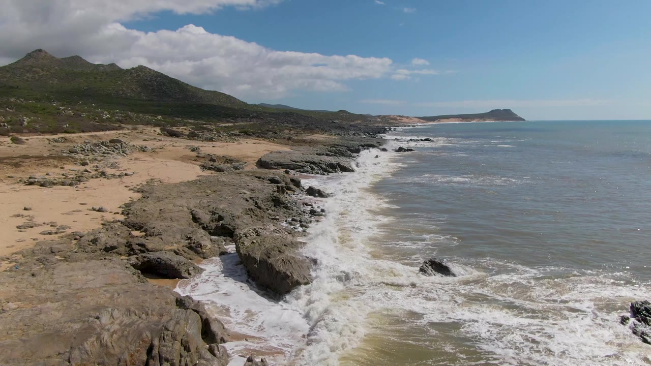 Buzzards Beach East Cape BCS Mexico frame with a large foreground rock ledge, whitewater spreading along the diagonal shoreline, muted blue ocean, distant headland, and no visible people.