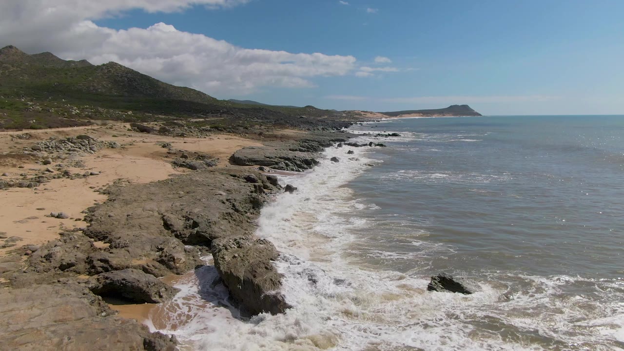 Buzzards Beach East Cape BCS Mexico frame showing the shoreline from slightly farther along the coast, with white foam trailing beside rocks, hills at left, open ocean at right, and no visible people.