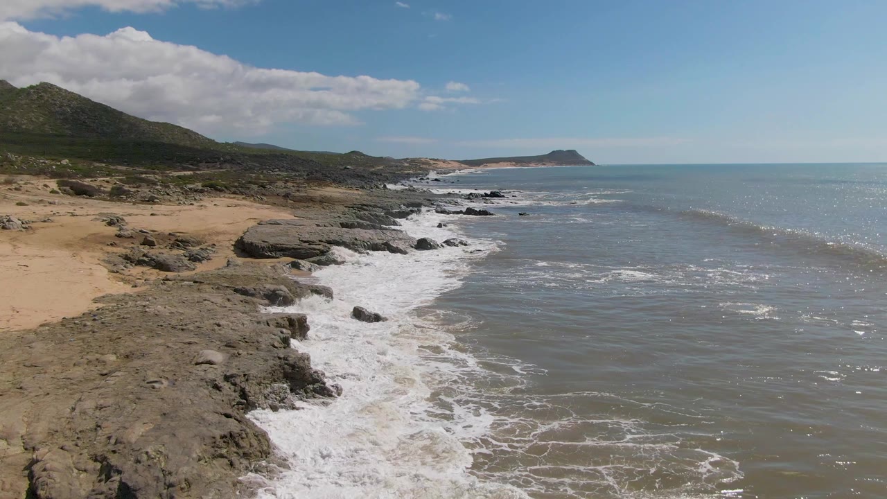 Buzzards Beach East Cape BCS Mexico shoreline with tan sand, dark rock shelves, white surf, green hills, blue ocean, and no visible people at the opening frame.