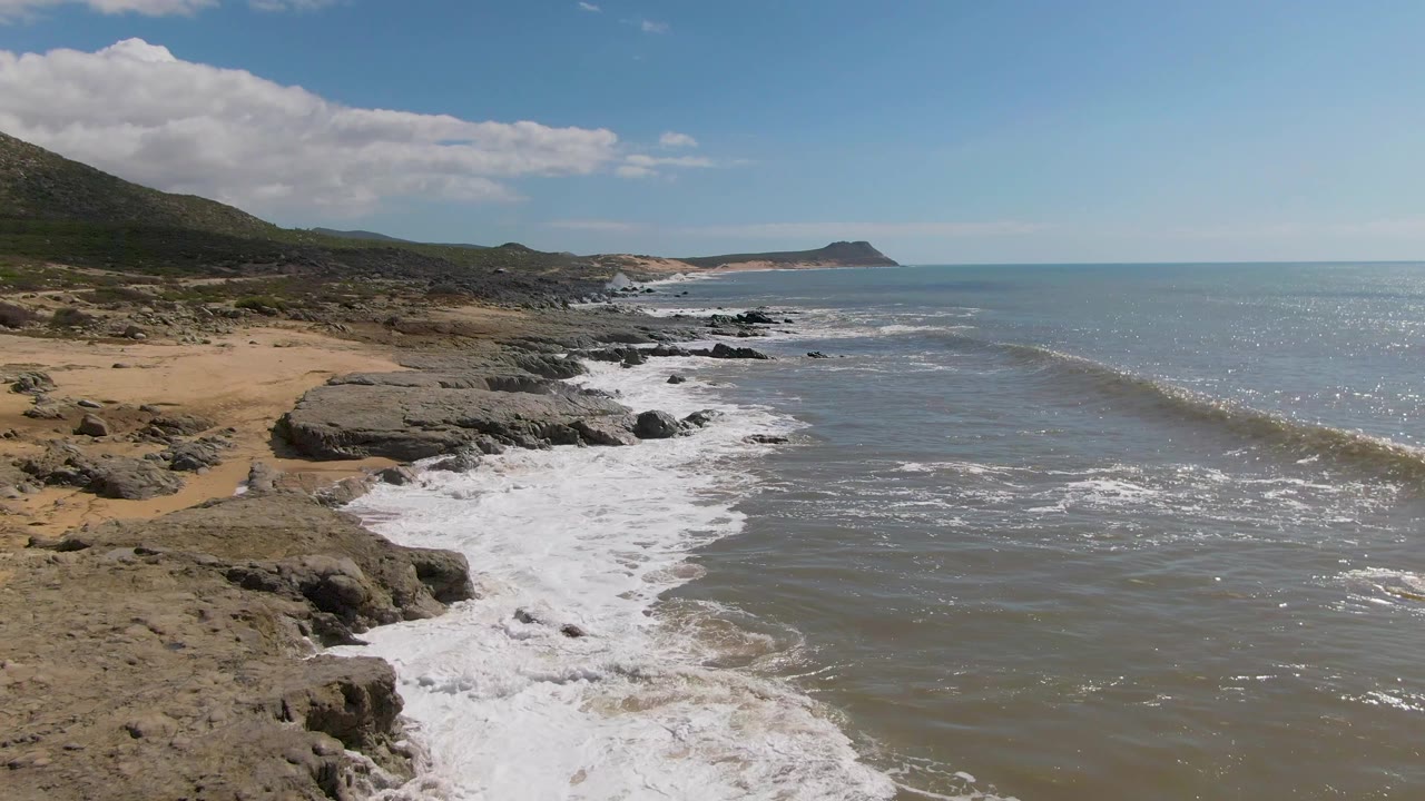 Buzzards Beach East Cape BCS Mexico coast with a small wave line forming offshore, white foam against dark rocks, low hills on the left, and no visible people.