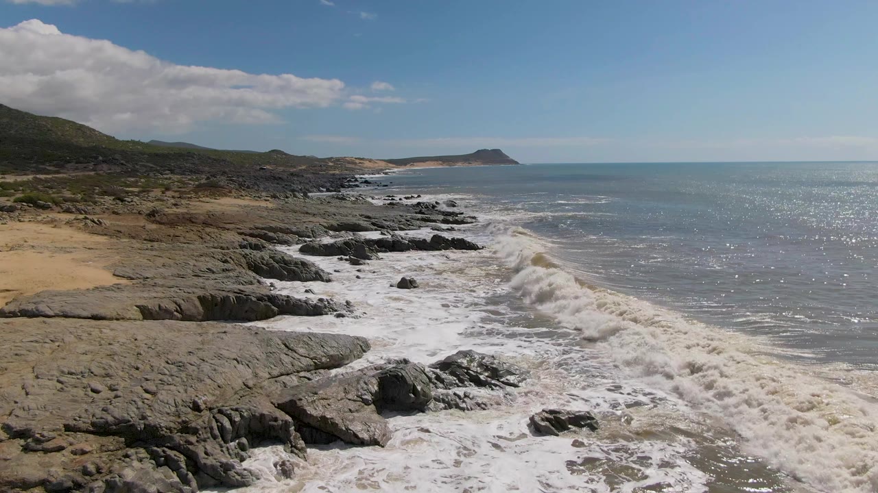 A foamy wave rolls along the Buzzards Beach rocky shoreline, with gray-blue ocean to the right, scrub-covered hills to the left, and no visible people.