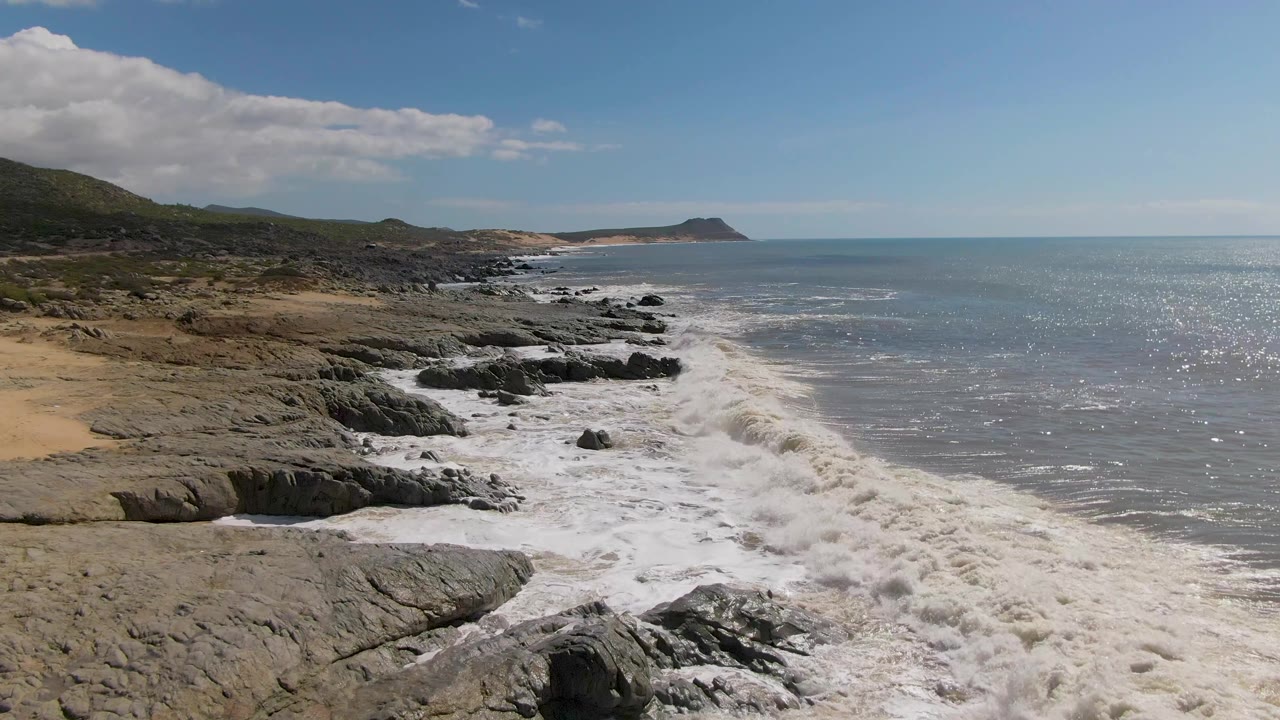 White water curls toward the rocks at Buzzards Beach East Cape BCS Mexico, while sun glare speckles the ocean and the sandy cove remains visible at left with no visible people.