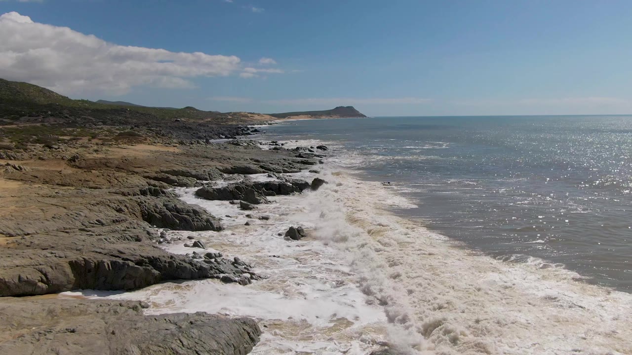 A wider elevated view shows the Buzzards Beach rock ledges running into foamy surf, with open water, distant hills, and no visible people in the frame.