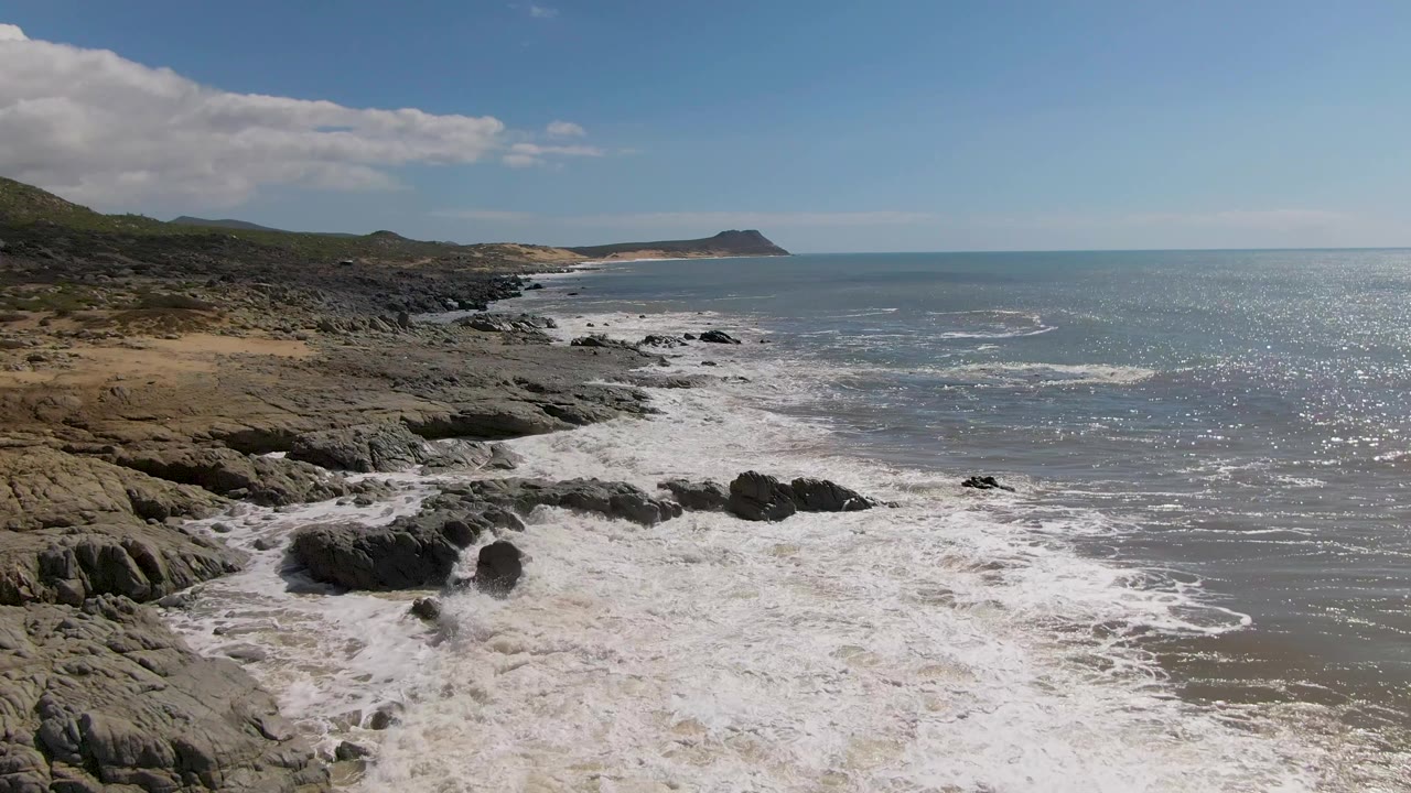First frame of Buzzards Beach East Cape BCS Mexico showing white surf hitting dark rocks, scrubby shoreline to the left, open ocean to the right, and no visible people.