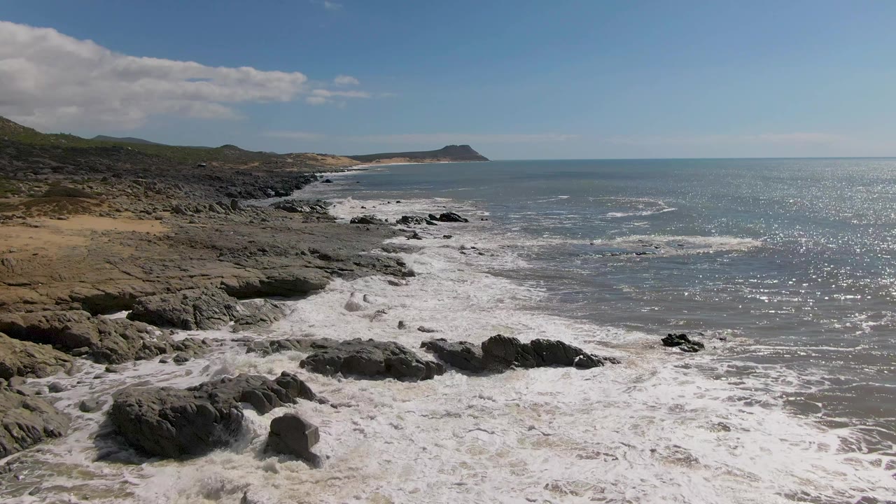 At 0:01, Buzzards Beach East Cape BCS Mexico is seen from an elevated angle as foam wraps around foreground rocks and the coastline continues into the distance with no visible people.