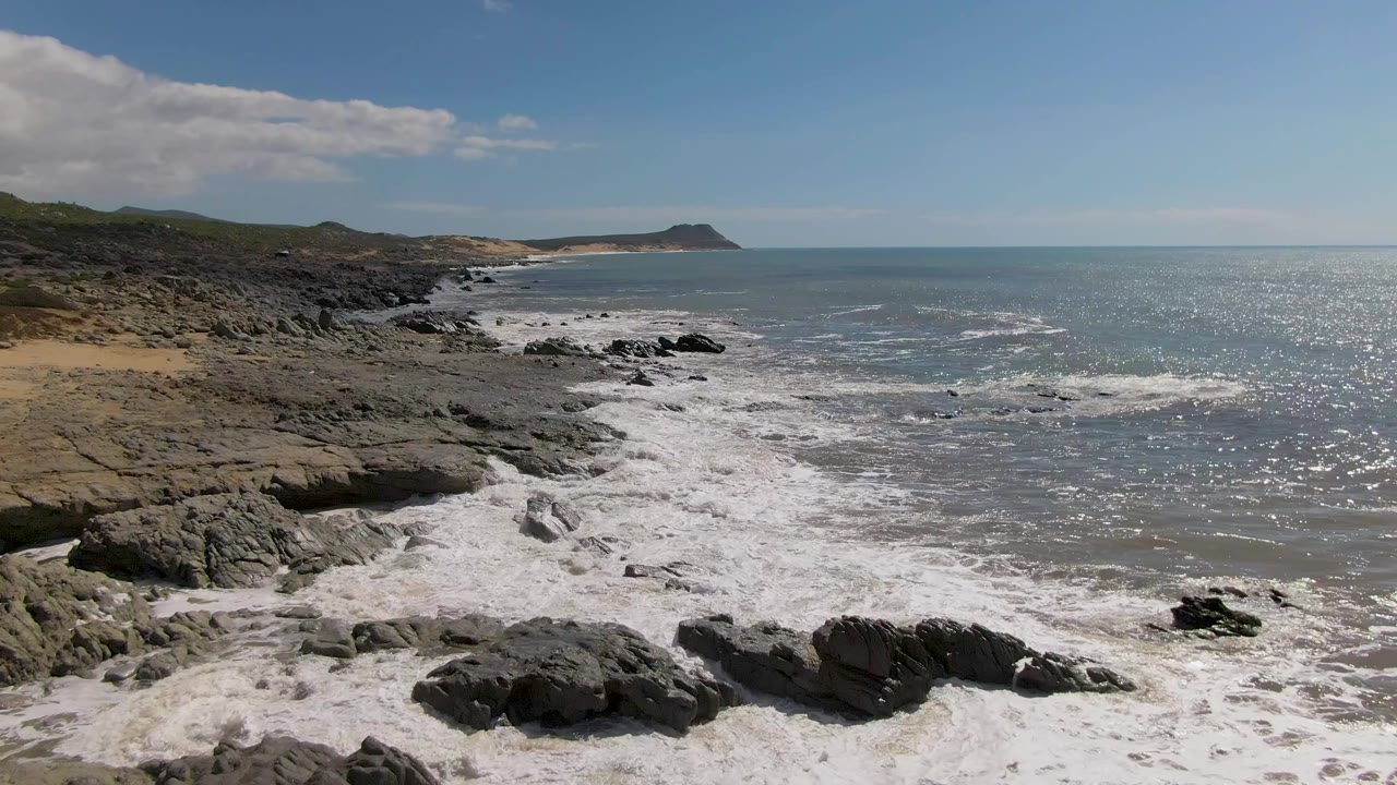 At 0:02, Buzzards Beach East Cape BCS Mexico shows a wider spread of whitewater across the foreground rocks, sun sparkle on the ocean, low hills at left, and no visible people.