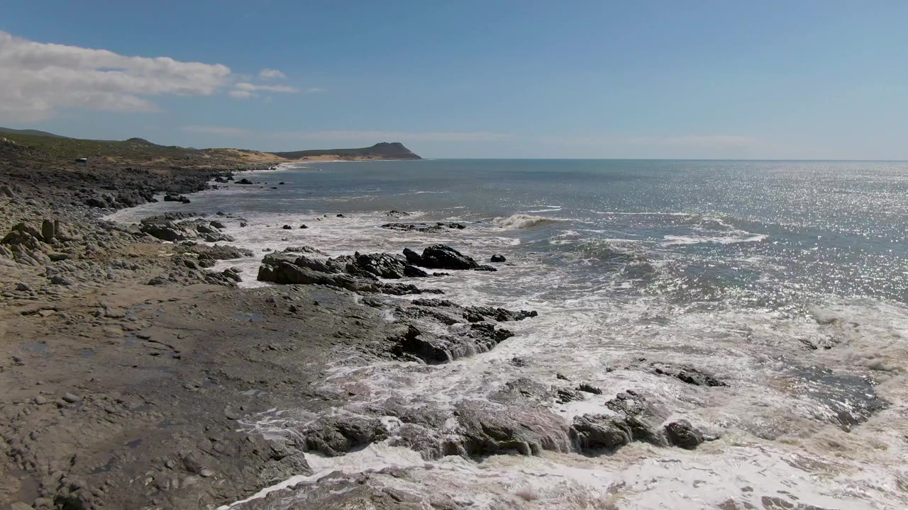 Elevated view of Buzzards Beach on Mexico's East Cape with white surf washing over dark rocks, a distant headland, scattered clouds, and no visible people.