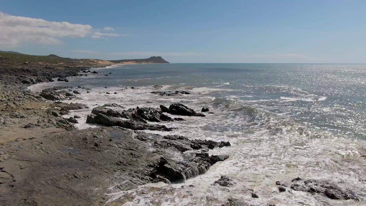 Buzzards Beach shoreline seen from above with foamy waves wrapping around black rock outcrops, low hills on the left, open ocean to the right, and no visible people.