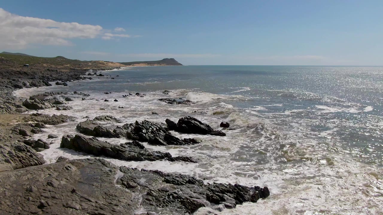 A rocky section of Buzzards Beach fills the foreground as churning whitewater pushes toward the camera, with a long ocean horizon and no visible people.