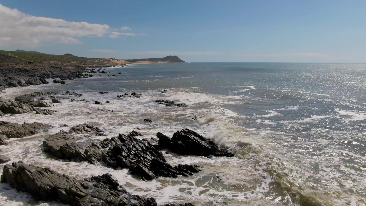 Wave foam surrounds jagged shoreline rocks near a distant headland.