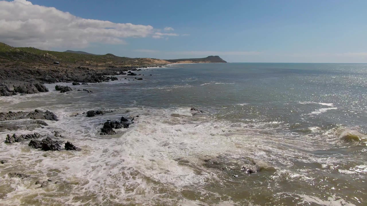 Rocky shoreline with white surf, green hills, open ocean, and partly cloudy sky.