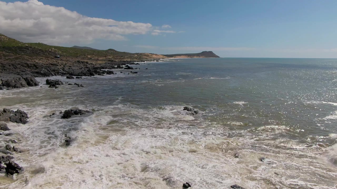 At Buzzards Beach on the East Cape of BCS Mexico, surf foam spreads farther around scattered black rocks while the hilly coastline recedes into the distance, with no visible people.