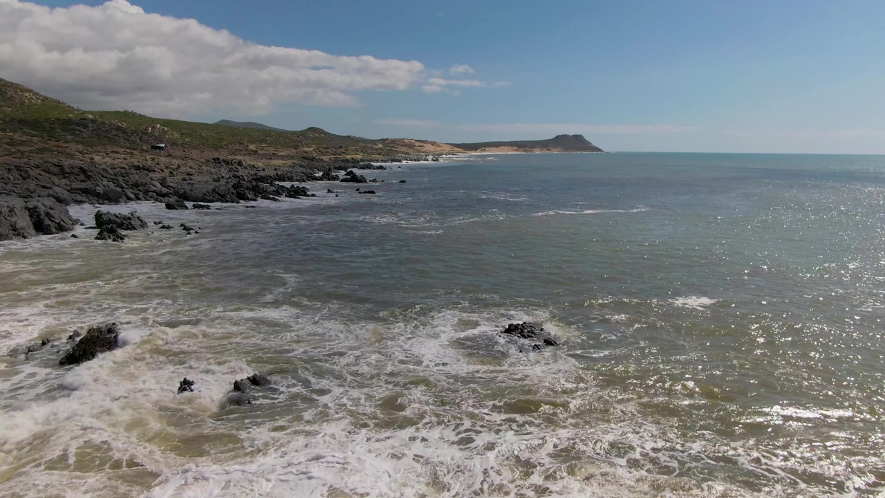 At Buzzards Beach on the East Cape of BCS Mexico, churning white water covers the near rocks as sunlight sparkles across the ocean toward a low headland, with no visible people.