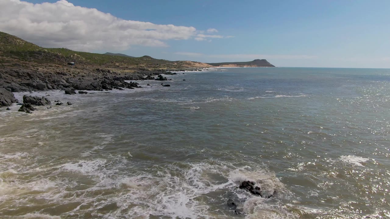 At Buzzards Beach on the East Cape of BCS Mexico, the surf has pulled back from several dark rocks, leaving more open water visible along the rocky coast, with no visible people.