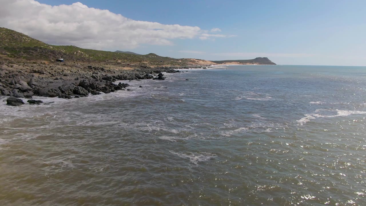 Rocky coast with waves, green hills, and partly cloudy sky.