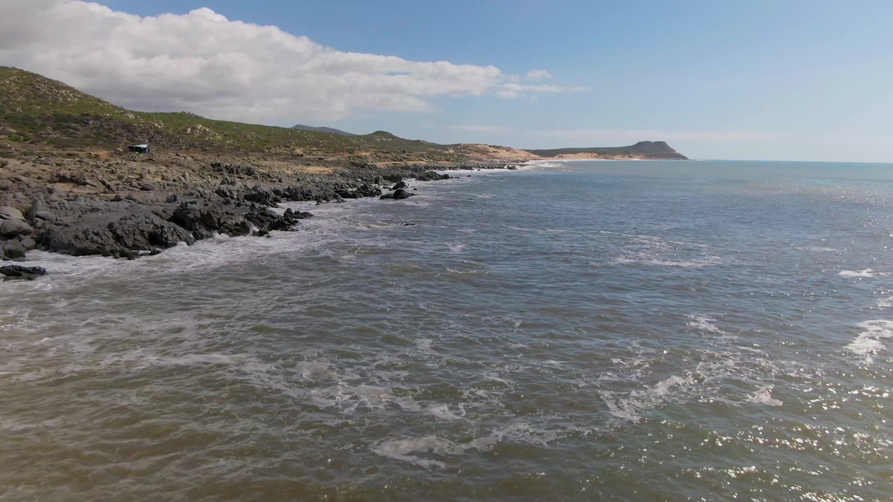Buzzards Beach East Cape BCS Mexico shoreline with foamy water spreading near the rocks, low hills along the left background, open sea to the right, and no visible people.