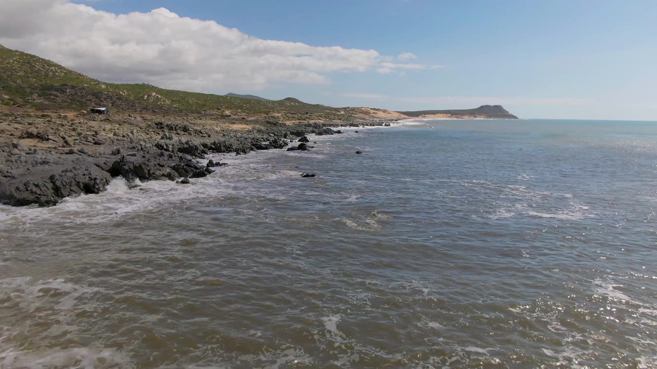 Mid-clip view at Buzzards Beach East Cape BCS Mexico with more open water visible on the right, scattered foam lines, rugged rocks on the left, and no visible people.