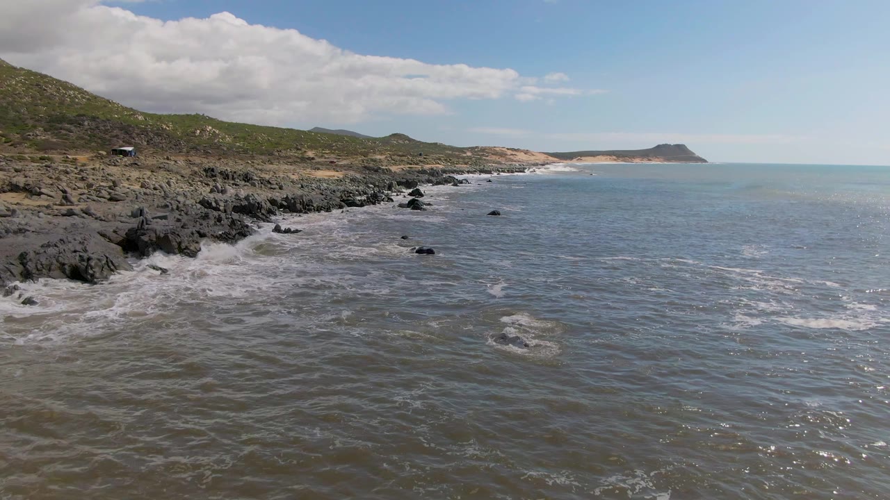 Buzzards Beach East Cape BCS Mexico frame showing choppy gray-blue water in the foreground, black shoreline rocks at left, sandy distant coast, and no visible people.