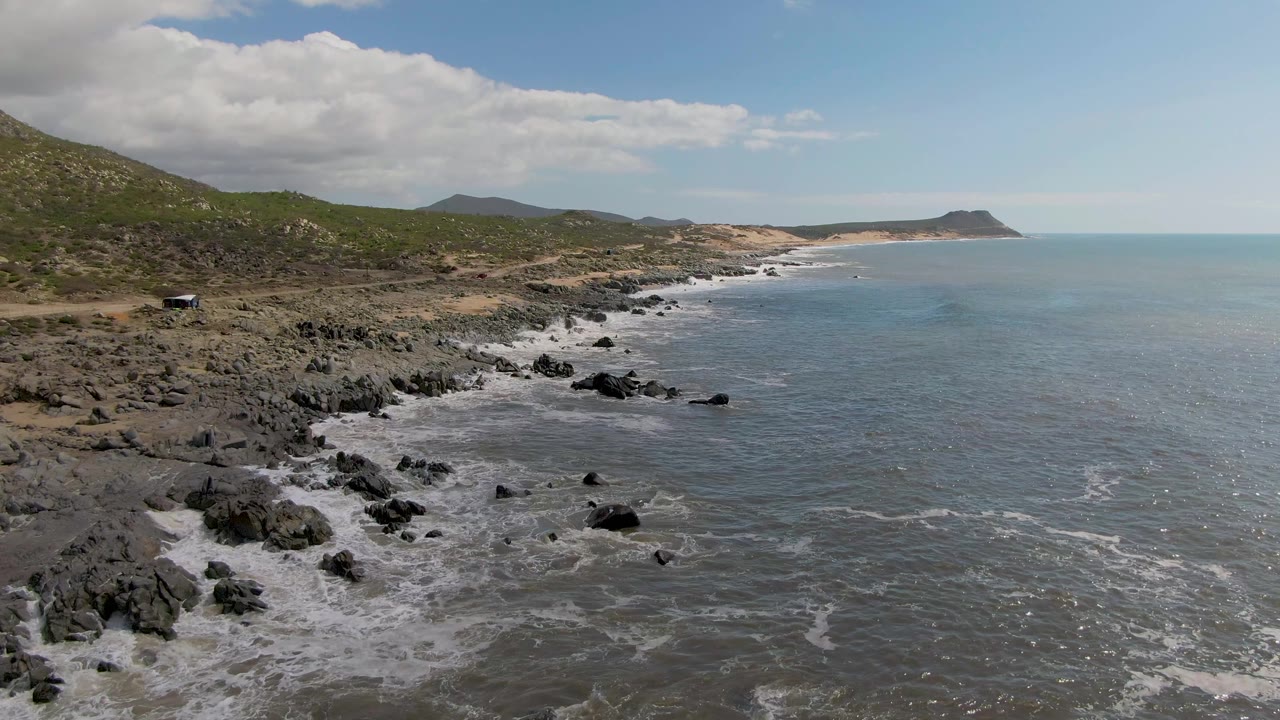 Aerial view of rocky coastline, ocean, green hills, and a small car on a coastal road.