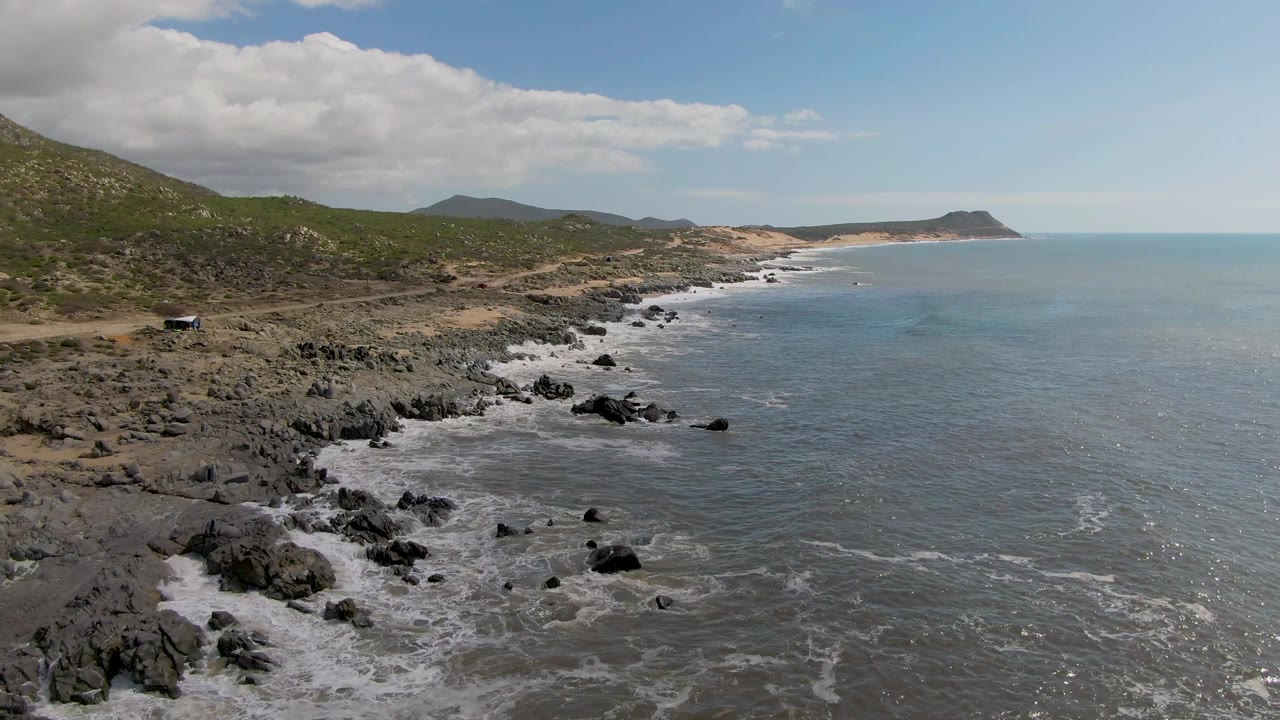 A second aerial view at Buzzards Beach East Cape BCS Mexico holds the small car along the hillside road while white waves spread around the dark rocks and no people are visible.