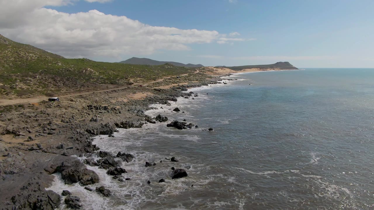 Aerial view at Buzzards Beach East Cape BCS Mexico with the car still visible on the road, foamy surf tracing the rocky edge, blue-green water to the right, distant hills, and no visible people.