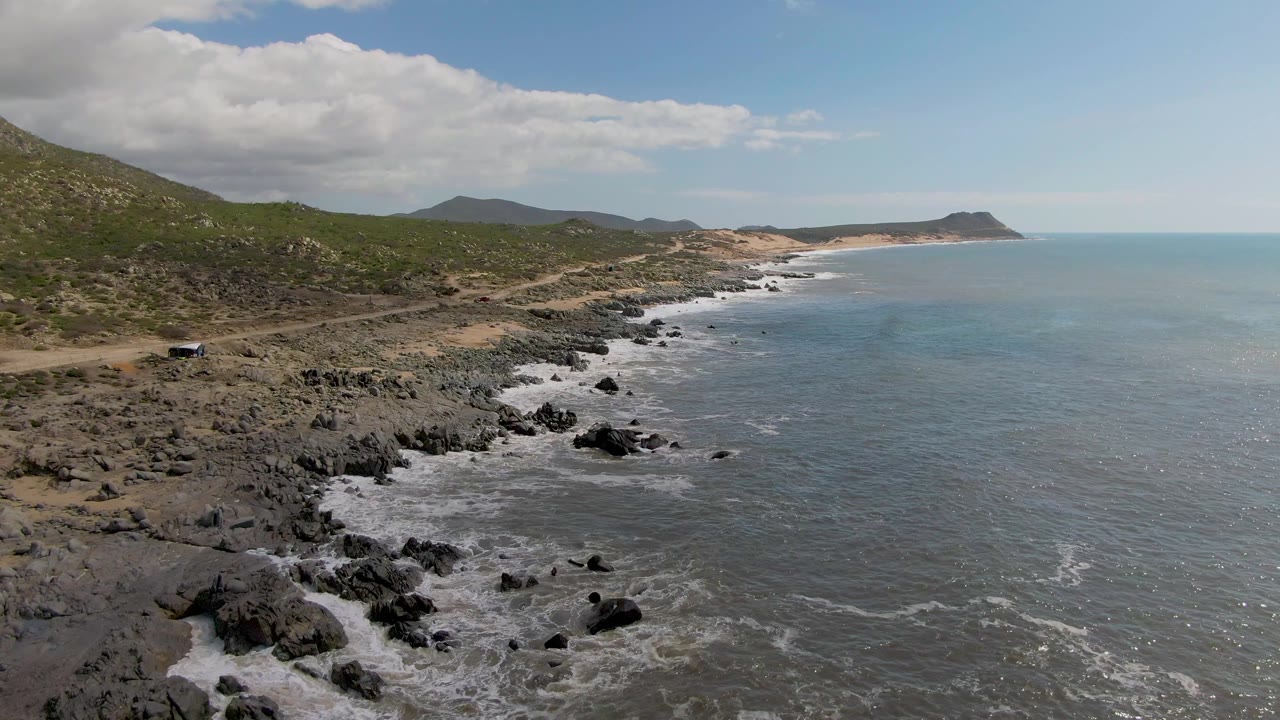 Mid-clip aerial frame at Buzzards Beach East Cape BCS Mexico showing a wider stretch of road and rocky coastline, the small car near the left side, low surf below, green hills behind, and no visible people.
