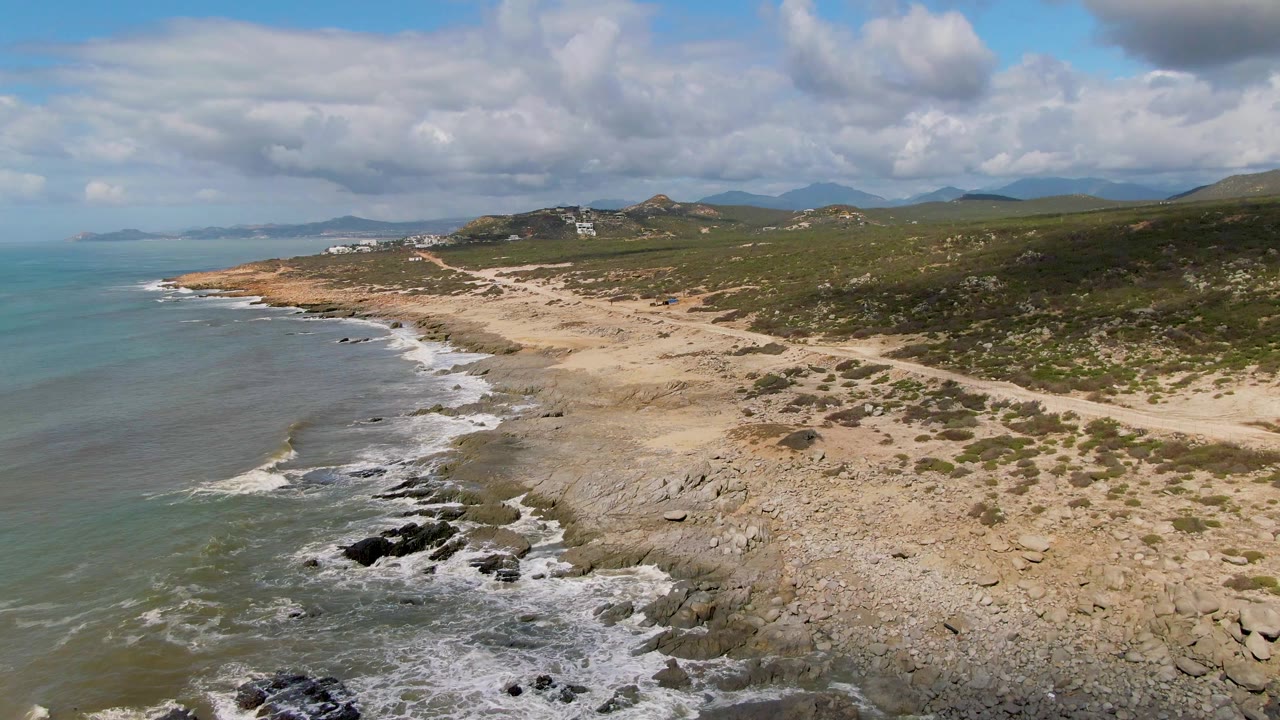 Aerial frame from Buzzards Beach East Cape BCS Mexico showing blue-green ocean, white surf on dark rocks, pale sand, scrub hills, distant buildings, cloud cover, and no visible people.