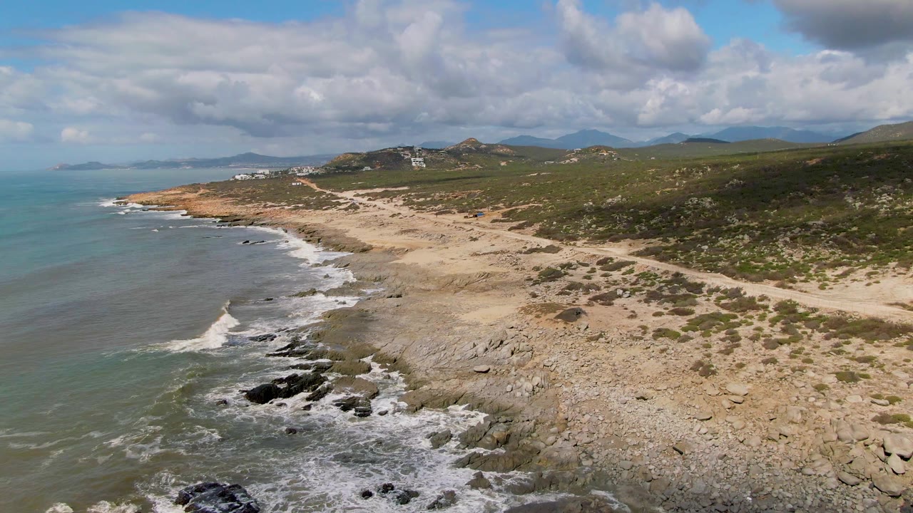 Drone view over Buzzards Beach East Cape BCS Mexico with surf pushing toward black rocks, a sandy and stony shoreline, green hillside terrain, distant structures, and no visible people.