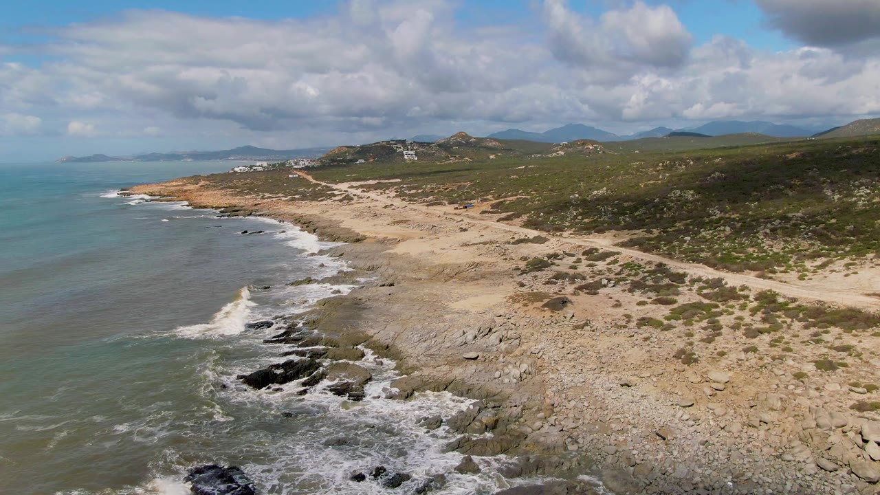 Aerial coastal view at Buzzards Beach East Cape BCS Mexico where a white wave breaks higher along the rock line, with ocean to the left, sandy shore to the right, hills beyond, and no visible people.