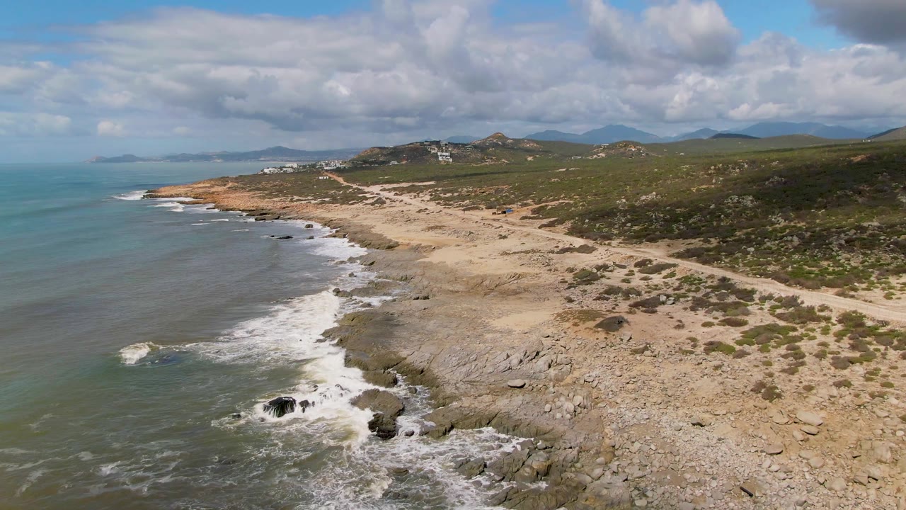 Aerial frame over Buzzards Beach East Cape BCS Mexico shows green-blue ocean, white surf on dark rocks, pale sandy shoreline, scrub hills, distant mountains, cloud cover, and no visible people.