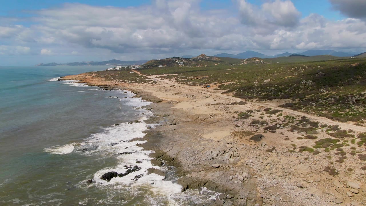 At 0:02, the East Cape coastline remains framed from above with waves curling into dark rocks, pale sand and stone in midframe, low green hills inland, and no visible people.